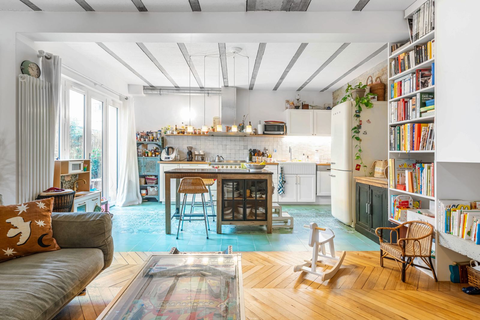 Le loft vu depuis le salon : poutres apparentes, îlot central, carreaux de ciment turquoise, parquet en point de Hongrie et bibliothèque